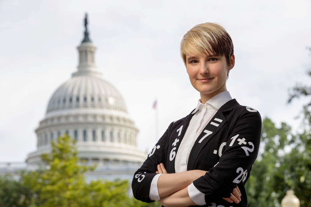 B poses with arms crossed in front of the US capitol donning a black jacket emblazoned with white mathematical symbols