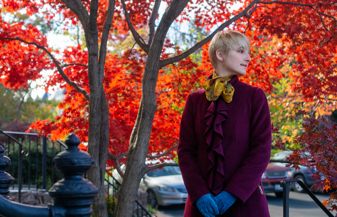 B stands amongst the vibrant red and orange foliage of fall in DC. They wear a maroon coat and blue gloves.