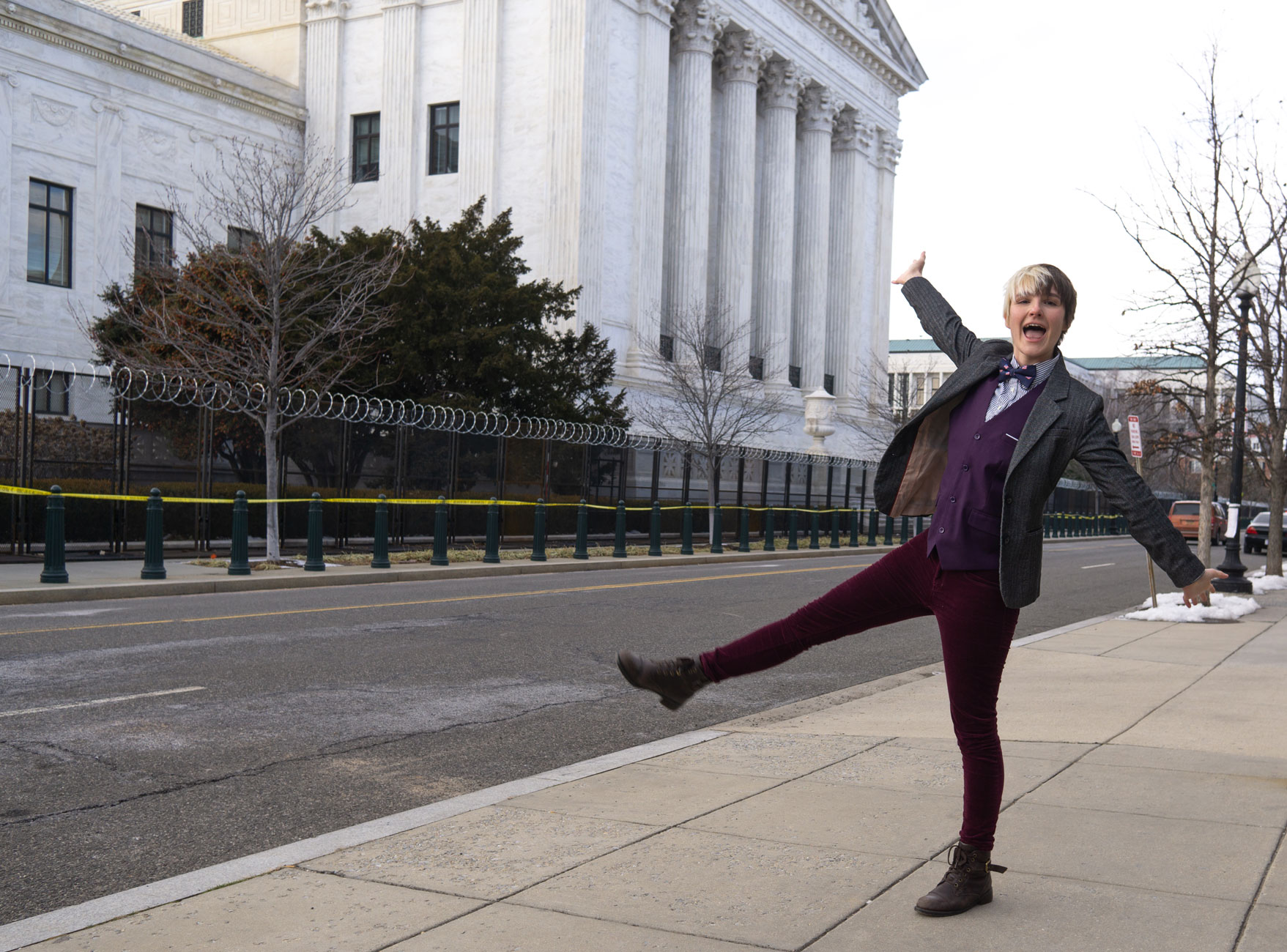 B kicks up a leg in an enthusiastic pose in front of the iconic columns of the Supreme Court, fenced in with the rest of the capitol compound with barbed wire and armed guards in response to the January 6th attack on the capitol.